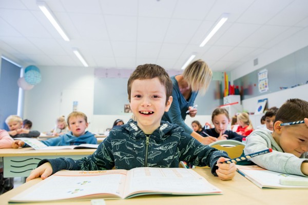 jongetje met kort haar en jasje met haaienprint aan lessenaar in de klas. Kijkt lachend in de lens met schrift voor zich en potlood in de hand.