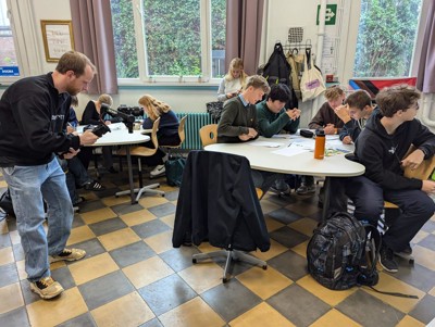 Een cameraman filmt in een klaslokaal waar leerlingen in groepjes aan een project werken