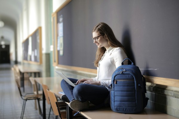 Vrouw zit op een tafel met een rugzak. Ze leunt tegen een schoolbord.